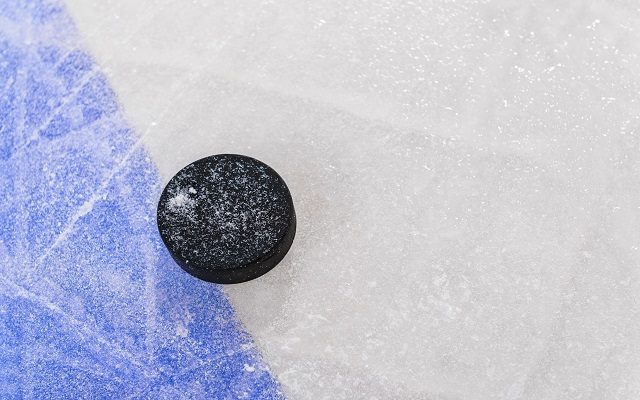 Looking down on a ice hockey puck sitting next to the Blue Line with marred up ice from the skate blades. The Blue Line separates the attacking and defending zones from the neutral zone in the center of the rink.